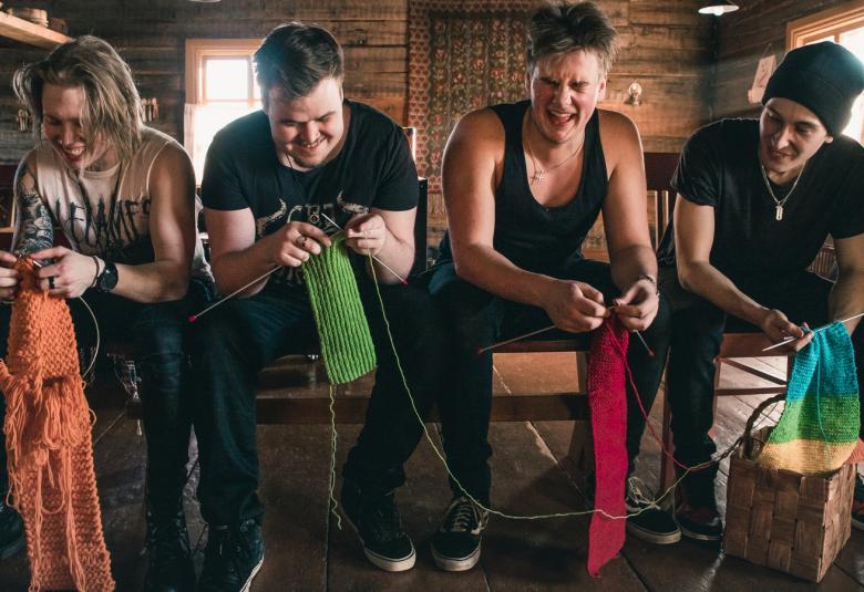 Kuvassa neljä nuorta miestä kutovat ja istuvat penkillä iloisin ilmein. Heillä on päällään mustat t-paidat ja yhdellä vaalean ruskea mustalla tekstillä.  Tila on vanha hirsinen huone, jossa on ryijy takaseinällä. Vasemmalla on ylhäällä hylly, jossa on keraamisia esineitä.