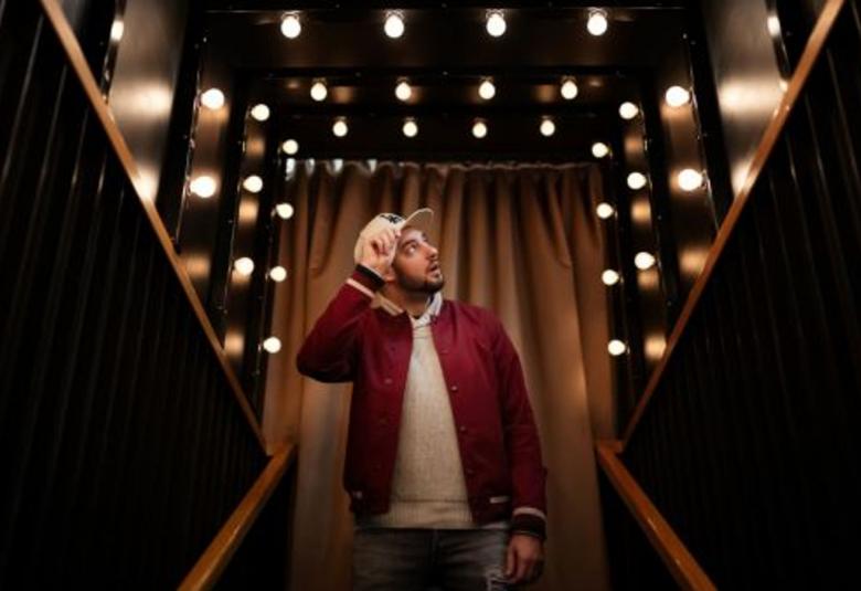 Kuvassa on puolivartalokuva HesaÄijästä, joka pitelee oikealla kädellään vaaleata lippalakkiaan ja katsoo vasemmalle ylös.  Päällään hänellä on tumman punaruskea pusakka ja alla vaalea paita.  Taustalla on valoja ja takana suoraan ruskea verho oviaukon päällä ja sivussa vinokaistaleet ruskeaa puuta.