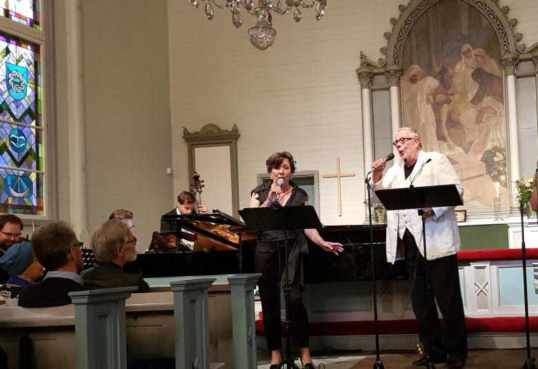 Kuvassa on vasemmalla Lilli Paasikivi ja hänellä on tummat hiukset ja musta housuasu.  Vieressä on Matti Salminen valkoisessa takissa ja mustissa housuissa.  Oikealla on Mari Palo kukallisessa mekossa, joka on puolisääreen. Hänellä on ruskeat hiukset.  Vasemmalla takana näkyvät viulisti Petteri Iivonen, Jukka Nykänen ja kontrabasisti Eero Ignatius. Etualalla näkyy yleisöä.