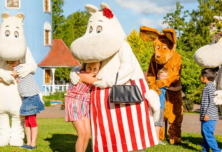 Vasemmalla on muumipappa ja keskellä muumimamma. Molemmat halaavat lapsia, jotka ovat paljon pienempiä kuin muumit.  Oikealla on muumi.  Takana näkyy sininen muumitalo.