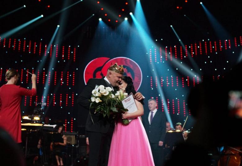 Kuvassa ovat tangokuninkaalliset 2019, vasemmalla Johannes Vatjus tummassa puvussa ja oikealla Pirita Niemenmaa vaaleanpunaisessa puvussa.  He halaavat toisiaan lavalla ja takana on punamusta tausta sekä sinisiä  valojuovia.  Vasemmalla on selin punaisessa mekossa oleva orkesterinjohtaja.