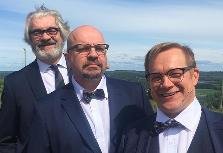 Kuvassa ovat Jussi Lampi, Timo Rautiainen ja Puntti Valtonen puolivartalokuvassa ja limittäin rinteessä.  Heillä on tummat puvuntakit päällä ja valkoiset paidat sekä tummat solmiot.  Keskellä olevalla Timo Rautiaisella on rusetti.  Taustalla on sininen, kirkas taivas.