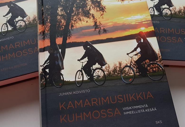 Kuvassa on kolme historiikkia, joiden kannessa on kuva kolme pyöräilevää soittajaan soittimet selässään.  Taustalla on järvi ja ilta-aurinkoinen taivas.  Kannessa lukee Kamarimusiikkia Kuhmossa, Viisikymmentä ihmeellistä kesää ja kirjoittaja Juhani Koivisto. Kuvat: Stefan Bremer. 