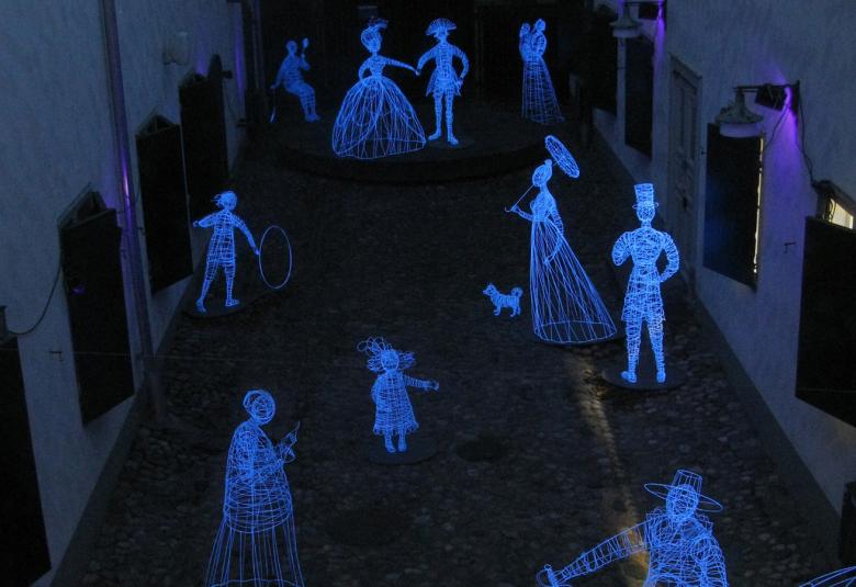 Kuvassa on tila joka on kahden seinän väli ja seinissä on ikkunat.  Tilan valo on tumma ja lattialla on valkoisia hahmoja koko tilassa.  Hahmot ovat miehiä ja naisia.  Vaatetus on vanhanaikaisen pönkkähameinen.