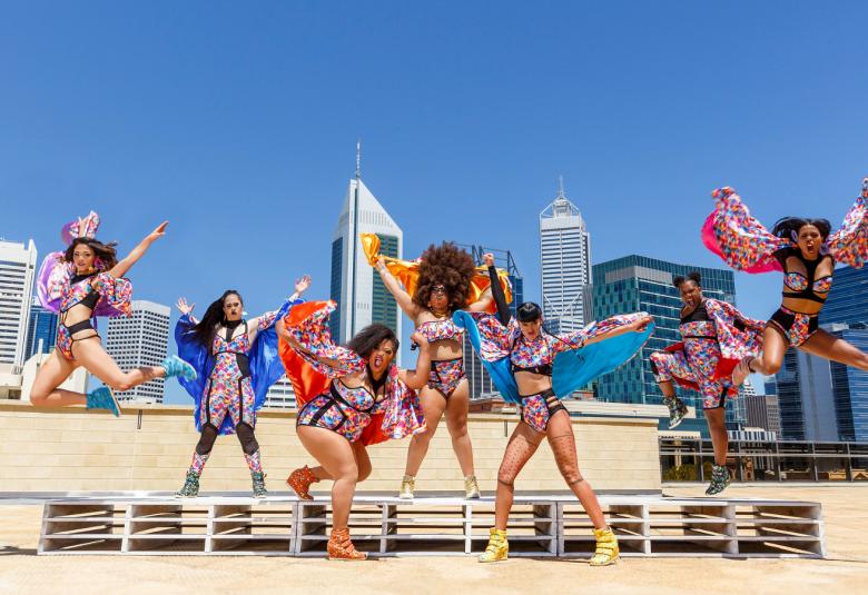 Kuvassa on Hot Brown Honey -tanssiryhmä puisella, vaalealla kattotasanteella ja takana on lasi-ikkunaisia, tummia ja vaaleita, korkeita toimistotaloja. Tanssijoilla on kirjavat esiintymisasusteet ja osalla ne ovat uimapukumaisia.  Taustalla on kirkkaan sininen taivas.