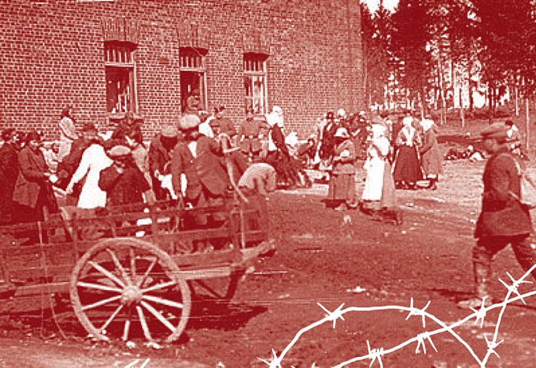 Kuva on ruskeasävyinen ja se kuvaa Hennalan vankileirin pihaa. Kuvassa on etualalla piikkilanka-aitaa ja tyhjät hevoskärryt.  Takana on vankeja, miehiä ja naisia.  Takana on tiilinen rakennus.  Takana oikealla on harvaa metsää.