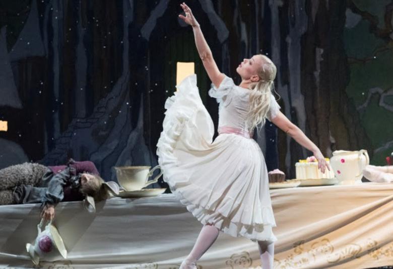 Kuvassa on Liisa Ihmemaassa baletin Liisa, Linda Haakana.  Hänellä on vaalea mekko ja hän osoittaa vasemmalla kädellään ylöspäin.  Lattialla makaa kani vasemmalla harmaassa puvussa.  Taustalla on tumman harmaa verho ja oikealla lattialla on kakkuja.