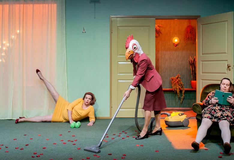 Kuvassa on kolme naista huoneessa.  Huoneessa on turkoosin harmaa lattia, vähn turkoosimpi seinä ja vasemmalla valkoisella ohuella verhottu ikkuna.  Kellertävät kaksoispuuovet oikealla ovat toisen oven kohdalta auki. Naisista vasemmalla on yksi lattialla kyljellään ja tekee jaloillaan v.kirjaimen.  Oven edessä on nainen murretun punainen jakku ja viininpunainen puolihame päällä imusoimassa lattiaa korkokengät jalassa.  Päässään hänellä on kukonpää.  Hänen oikealla puolellaan oven edellä rönottää nainen noja