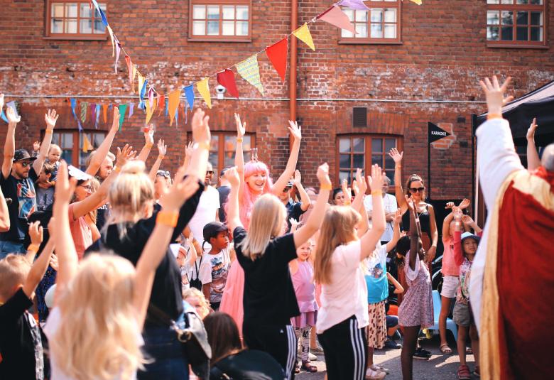 Kuvassa on Skidit festarit vuonna 2018 ja lapsia koko kuvan alueella kädet pystyssä.  Oikealla on reunassa yksi aikuinen ruskeassa viitassa kädet ylhäällä.  Taustalla näkyy rakennuksen tiiliseinää.