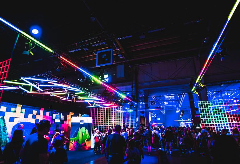 Kuvassa on Tubeconin kävijöitä.  Kuva on sävyltään tumma ja keskellä on värikkäitä valoja ja valokiilat kuvan kumpaankin kulmaan. Kuvan keskellä näkyy metalliruuturakenteita, joita on valaistu vaaleanvihreillä ja punaisilla valoilla ja tausta on sininen.  Samassa kohdassa näkyy kävijöitä selin.