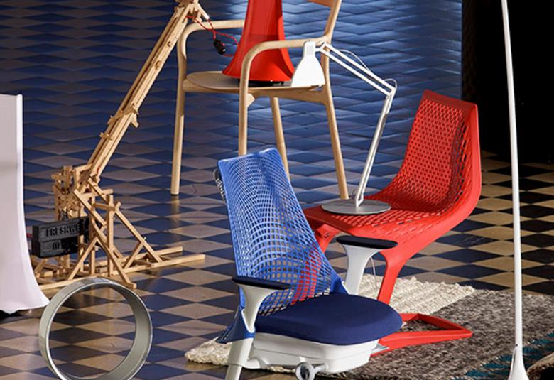 Kuvassa on ruskea-mustaruudullisella lattialla kaksi toimistotuolia, joista toinen on sinireiällisellä selkämyksellä varustettu ja toinen on punainen toimistotuoli. Lisäksi näkyy puista lampun jalkaa ja valkoinen pöytälamppu. 