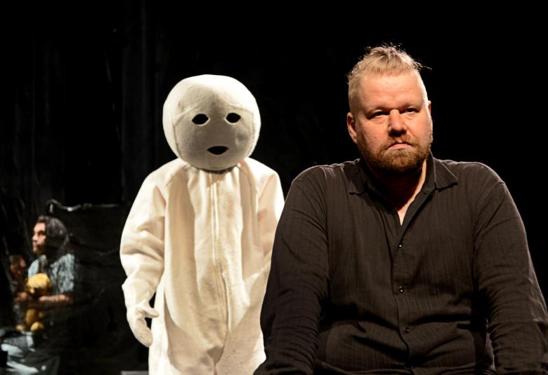 Kuvassa on mieshenkilö oikealla, joka istuu kuvan oikealla puolella ja on mustassa paidassa.  Miehellä on partaa ja päälaella vähän solmuun vedettyä tukkaa.  Takana on valkoinen pyöreäpäinen ja valkoisessa takissa oleva hahmo.  Tausta on musta ja kauempana vasemmalla takana istuu mies istumassa lattialla.