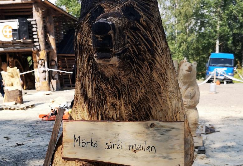 Kuva on on puusta tehdystä karhusta, jonka etuosassa on kyltty Mörkö särki mailan.  Karhun pää on tummaraitainen.  Taustalla näkyy vasemmalla hirsimökki ja oikealla sininen pakettiauto koivikon edessä.