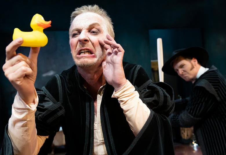 Kuvassa on Eero Aho Don Juanina ja hänellä on päällään musta viitta.  Aholla on kasvoillaan irvistys ja hänellä on kädessään keltainen pieni muoviankka, jota katsoo. Takana näkyy toinen henkilö, joka on myös mustassa viitassa ja katsoo Ahoa.  Tausta on musta.