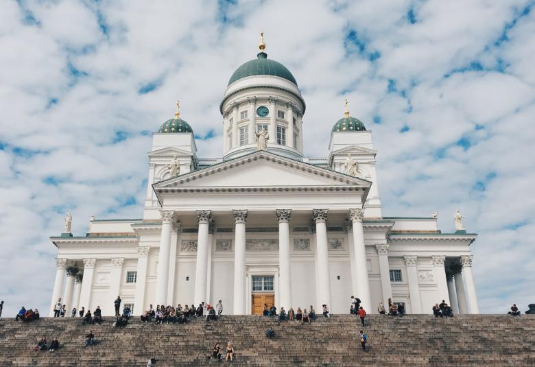 Kuvassa on Helsingin tuomiokirkko, joka on väriltään valkoinen.  Taustalla on sinistä taivasta ja siinä on valkoisia pieniä pilviä.  Alempana on ylempiä portaita ja niillä istuu ihmisiä siellä täällä.
