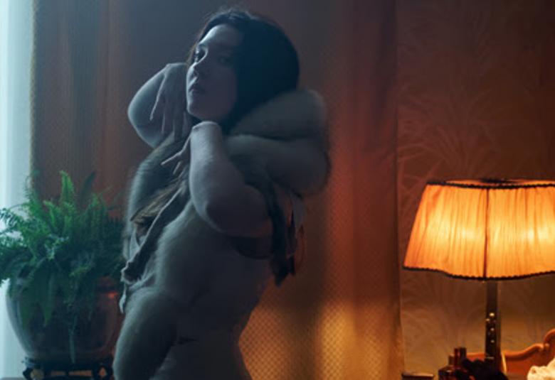 Kuvassa on nuori nainen harmaan värisenä turkiskaulus kaulassaan.  Kuva on huoneesta, jossa on ruskea verho takana ja vasemmalla valkoista verhoa sekä edessä saniainen.  Oikealla on ruskehtava pöytälamppu, jossa on valo.