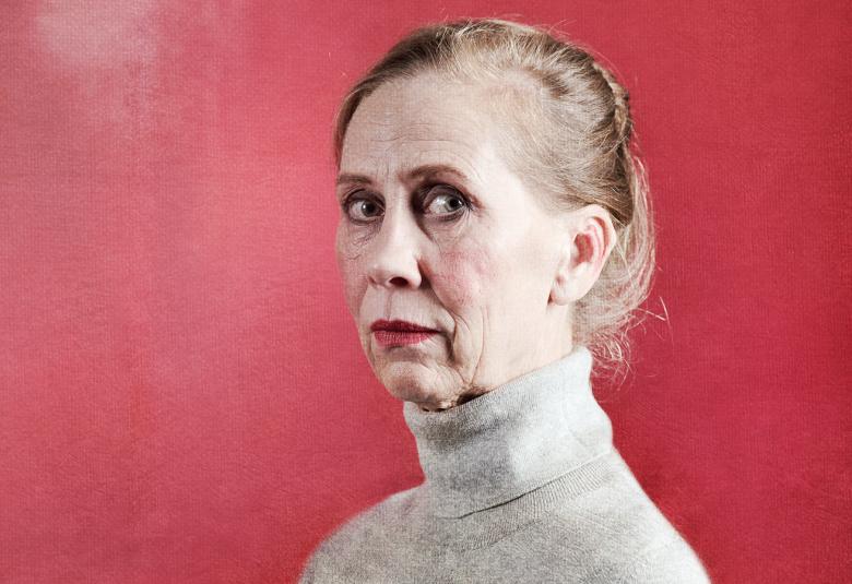 Kuvassa on Kati Outinen ja hänellä on vaalean harmaa poolopusero päällään.  Kuva on puolivartalokuva.  Outisella on ruskeat taakse vedetyt hiukset ja hän katsoo kameraan. Kuvan tausta on korallin punainen.