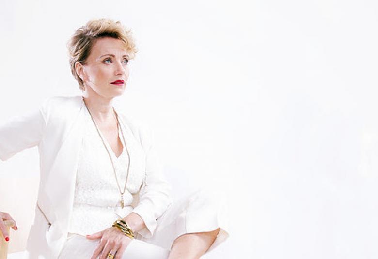 Kuvassa on vaalealla pohjalla valkoisessa jakkupuvussa Susanna Airaksinen. Jakkupuvussa on myös valkoinen liivi.  Airaksisella on lyhyet vaaleat hiukset. Airaksinen istuu vinottain tuolilla.  