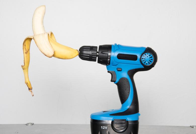 Kuvassa on sini-musta pora, jonka poranterässä on avattu banaani.  Pora on jalustallaan harmaalla teräspinnalla ja kuvan tausta on vaalean harmaa. 