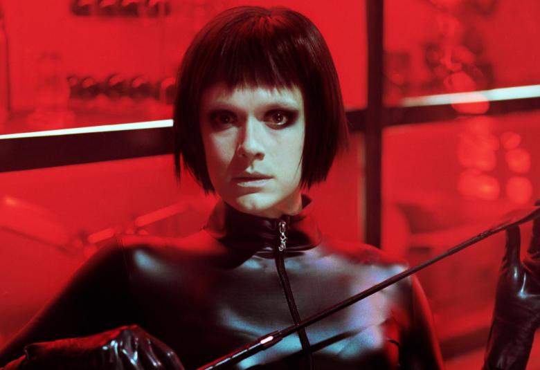 Kuvassa on Krista Kosonen domina-asussa mustassa korkeakauluksisessa nahkapuserossa, jossa on vetoketju edessä. Kososella on kädessään piiska.  Hänellä on musta polkkatukka ja tumma silmämeikki.