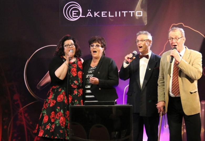 Kuvassa on Eläkeliiton karaokemestaruuskilpailut 2018.  Kuvassa ovat voittajat oikealta Kaija Pietilä, Arja-Inkeri Joas, Oiva Makkonen ja Jukka Korpela. Pietilällä on punakukallinen, mustapohjainen mekko, Joaksella musta bleiseri ja mustavalkoraidallinen pusero. Oiva Makkosella on musta puku ja valkoinen paita sekä tumma rusetti.  Korpelalla on vaalean ruskea takki, valkoinen paita ja mustat housut sekä puna-raidallinen kravatti. Kaikki pitävät mikrofonia kädessään. 