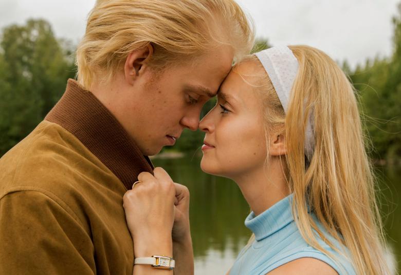 Kuvassa on Tatu Sinisalo ja Talvikki Eerola puolivartalokuvassa ja he ovat kasvot vastakkain.  Sinisalolla on vaalean ruskeat hiukset ja ruskea pusakka.  Eerolalla on vaaleat pitkät hiukset ja valkoinen kangaspanta päässä sekä hihaton vaaleansininen hihaton pusero päällään.  Taustalla on kesäistä järvimaisemaa.