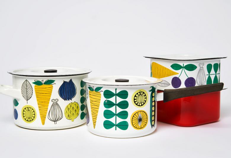 Kuvassa on kaksi kattilaa vasemmalla ja niissä on valkealla pohjalla selkeitä pystysuuntaisia, graafisia porkkanoita ja muiden vihannesten kuvia.  Oikealla on neliskulmaiset kupit, joista toinen on punainen ja toinen vihanneskuvioitu