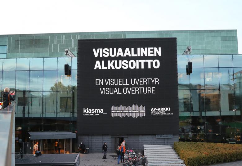 Kuva on Musiikkitalosta harmaana päivänä.  Musiikkitalon isolla näytöllä lukee Visuaalinen alkusoitto, en visuell uvertyre,visual overture. Alla on yhteistyökumppaneiden Kiasma, Helsingin kaupunginorkesterin ja Av-arkin logot.