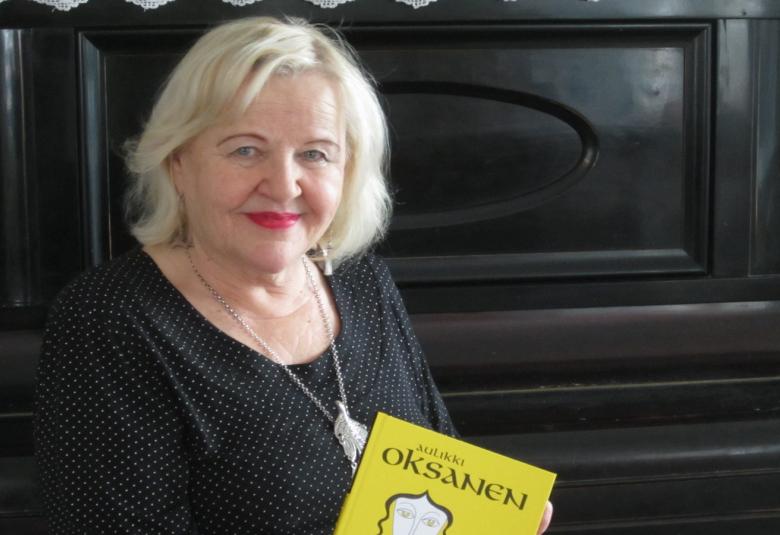 Kuvassa on Aulikki Oksanen ja hänellä on vaaleat hiukset ja musta valkopilkullinen mekko. Hänellä on kädessään keltainen, kirjoittamansa kirja.  Tausta on tummapuinen.