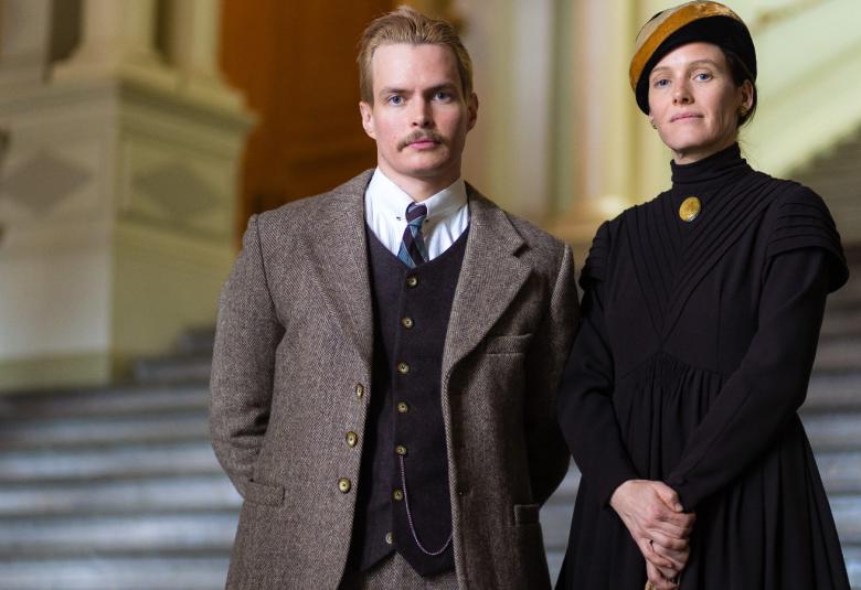 Kuvassa on vasemmalla Laura Birn ja oikealla Johannes Holopainen.  Molemmat ovat vanhanaikaisissa vaatteissa. Taustalla on vaalea portaikko yhdesssä Senaatintorin rakennuksista.