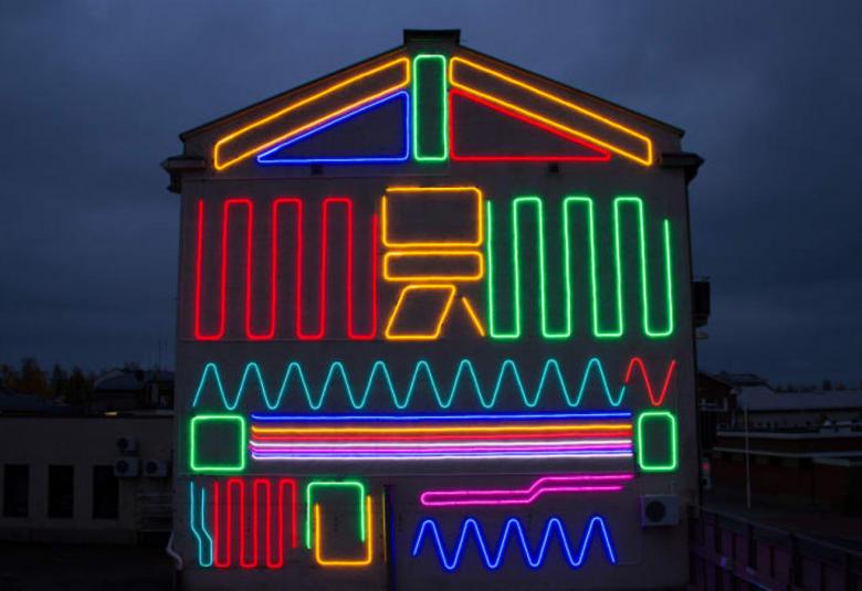 Kuvassa on Iisalmen kaupungin Neon Mural#6, jossa on tummassa iltavalossa kuvattu talon pääty.  Siinä on valojuovia, jotka on ovat eri värisiä.  Päätykolmiossa on vihreä ja punainen kolmio sekä keltainen palkki.  Keskella on keltaisia kuvioita ja vihreä ylös-alas menevä juova.  Alempana on sinistä sahalaitaa ja punaisia sekä liloja vaakaviivoja. alhaalla on punaista ja sinistä sahalaitakuviota ja vihreät ja keltaiset u-kuviot.