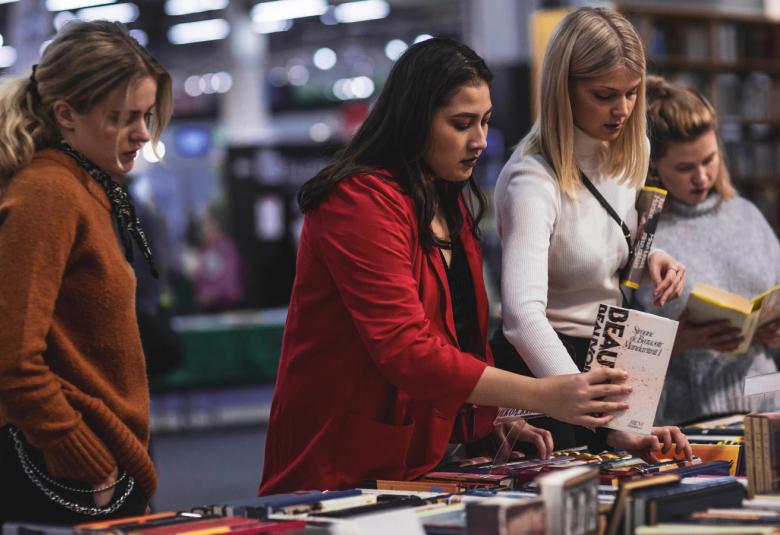 Kuvassa on kolme nuorta naista katsomassa kirjoja Helsingin Kirjamessuilla. Heidän edessään on täysi kirjapöytä.