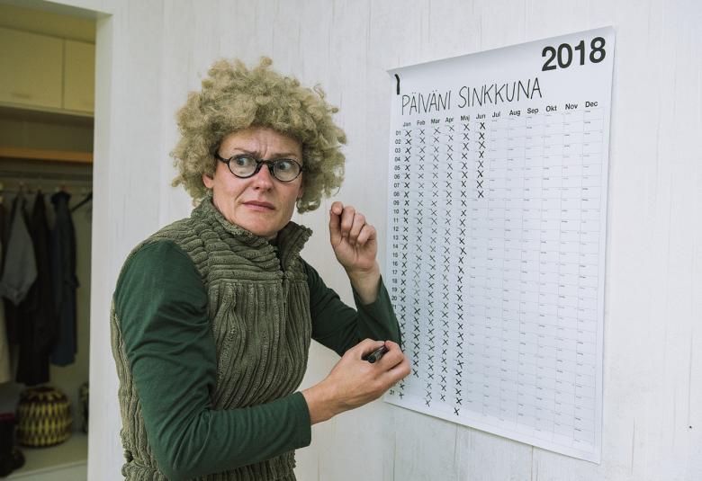 Kuvassa Pirjo Lonka, joka esittää Krapula-Päiviä. Lonka on keskellä kuvaa vihreässä samettipuvussa ja vaaleahkossa kiharaperuukissa sekä silmälaseissa.  Longalla on tussi oikeassa kädessään, jolla on rastii seinällä olevasan Päiväni sinkkuna 2018 kalenteriin rasteja.  Taulukossa on jo noin puolet rasteja.  Takana näkyy kaapissa takkeja ja kengät.