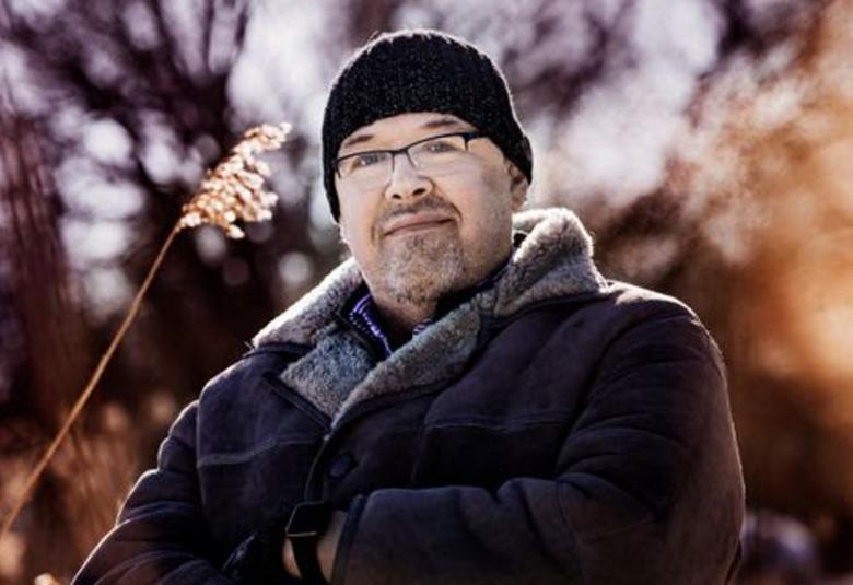 Kuvassa on Marcus Rosenlund, jolla on musta pipo päässään ja silmälasit.  Lisäksi hänellä on hiukan leukapartaa ja viikset.  Hänellä on tumman ruskea kelsiturkki päällään ja siitä näkyy kauluksessa harmaata karvaa.  Tuastalla on epätarkan näköistä metsää ja sävy on ruskea ja oikealla on valoläiskä.