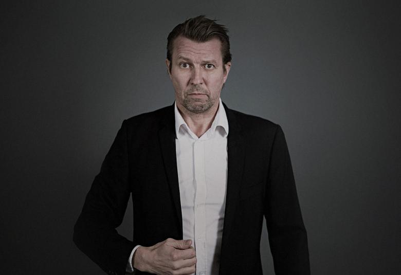 Kuvassa on Martti Suosalo, joka on vakavana musta puvun takki ja valkoinen paita päällä.  Tausta on harmaan tumma.