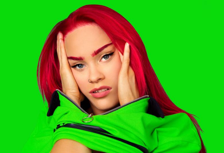 Kuvassa on Sanni, jolla on punaiset pitkät hiukset ja vihreä pusero.  Tausta on samaa vihreää kuin pusero. Sannin kädet ovat poskien päällä.    