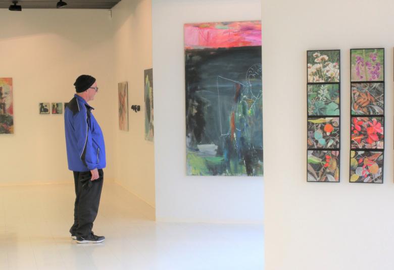 Kuvassa on Riihisaaren museon näyttely tila ja siinä ripustettuja näyttelyn tauluja eri seinillä.  Näyttelyä katsomassa on sivuttain sinisessä tuulipuvussa oleva mies.