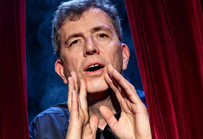 Kuvassa on Joachim Wigelius, joka kurkistaa punaisten samettiverhojen raosta.  Hänellä on kädet edessä kuiskausasennossa.  Witeliuksella on lyhyet, ruskeat hiukset ja sininen paita.  Taustalta näkyy tumman sinistä.