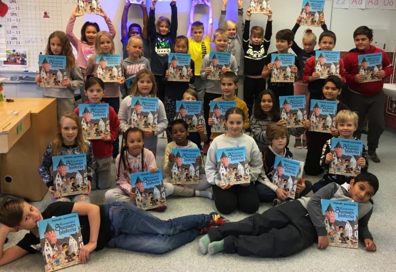 Kuvassa on koululaisia, joilla on Koiramäen historia kädessään.  Lapset ovat luokkakuva-asetelmassa.  Lapset ovat eri etnisistä ryhmistä ja pukeutuneet eri värisiin paitoihin ja housuihin.  Takana oikealla näkyy lavuaari ja vasemmalla on pulpetteja.   Lattia on vaalea.