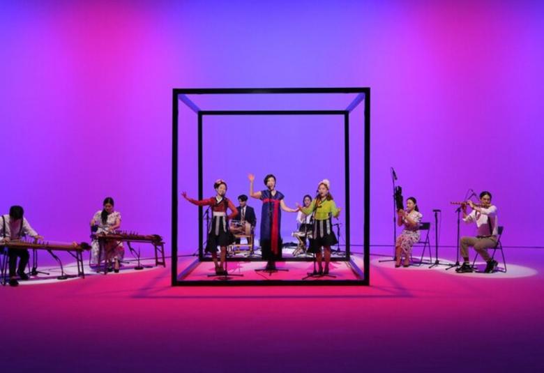 Kuvassa on sinisellä taustalla etelä-korealainen musiikkiryhmä.  Viisi esiintyjistä on mustareunaisen läpinäkyvän kuution sisällä.  Kaksi soittajaa, joilla on vaakasuorat, pöytätyyppiset soittimet on vasemmalla ja kaksi soittajaa ylöspäin suuntautuvilla soittimilla on oikealla puolella.