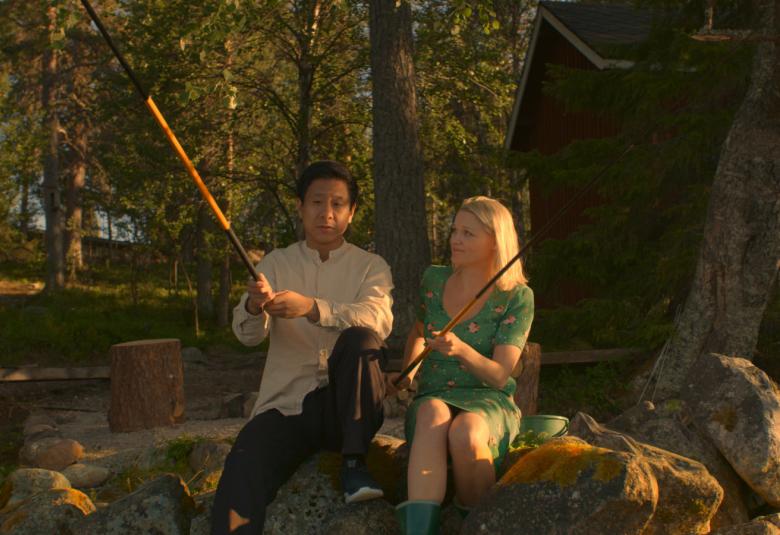 Kuvassa ovat Chu Pak Hong ja Anna-Maija Tuokko ongella Ounasjoen varrella. Rannalla on kivikkoa ja taustalla mänty-kuusimetsää ja punavalkoinen mökki. 