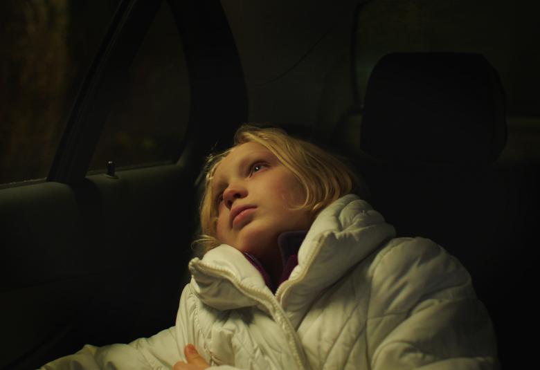 Kuvassa on nuori Helena Zengel, joka makaa puoliksi auton takapenkillä ja katsoo autosta ulos.  Hänellä vaaleat puolipitkät hiukset ja päällään valkoinen toppatakki.  Tausta on tumma.   