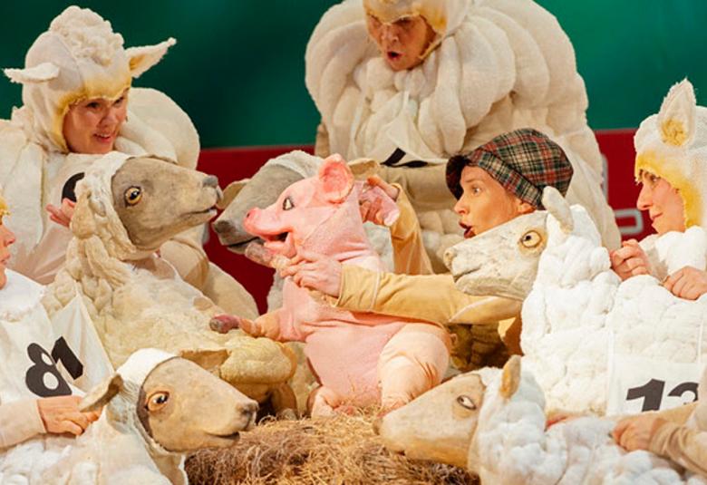 Kuvassa on näyttelijöitä lampaiden vaatteissa ja keskellä on Babe-possu nukkena ja muutama lammas.  Lampaat ovat valkoisia ja Babe vaaleanpunainen.