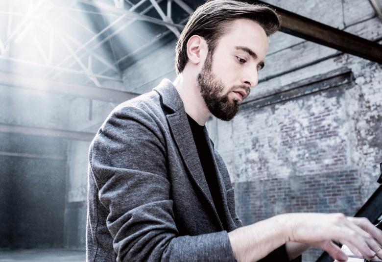 Kuvassa on Daniil Trifonov soittamassa pianoa harmaassa takissa. Kuva on harmaa sävytteinen.