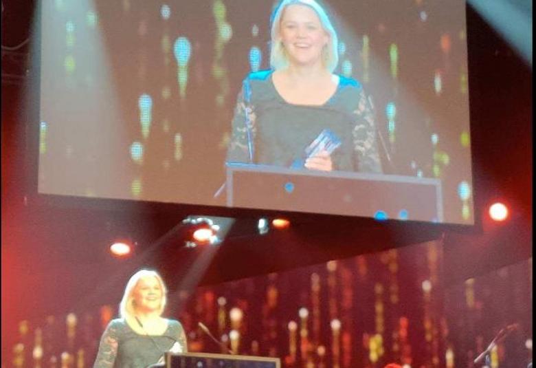 Kuvassa on Anna-Maija Tuokko pitämässä puhetta glitter-mekossa ja taustalla on monitori, josta näkyy Tuokon kuva.  Kuva on näyttämöltä.