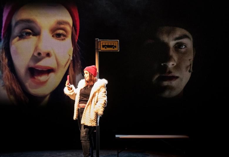 Kuvassa on nuori nainen bussipysäkillä ja katsoo kännykkäänsä.  Taustalla näkyy omat kasvot ja nuoren miehien kasvot.