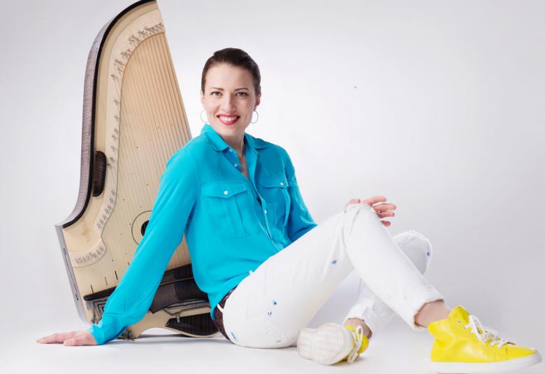 Kuvassa on Ida Elina kanteleen kanssa. Hän istuu lattialla ja kantele on hänen selkänsä takana.  Ida Elinalla on turkoosi paitapusero ja valkoiset farkut sekä keltaiset tennarit.