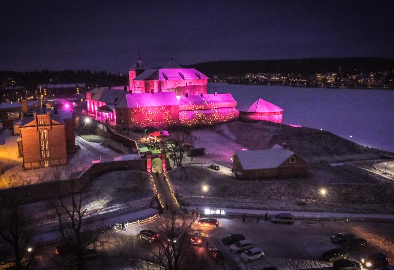 Kuvassa on Hämeen linna valaistuna punertavaksi ympäristössään.  Kuva on otettu illalla ja pimeällä.