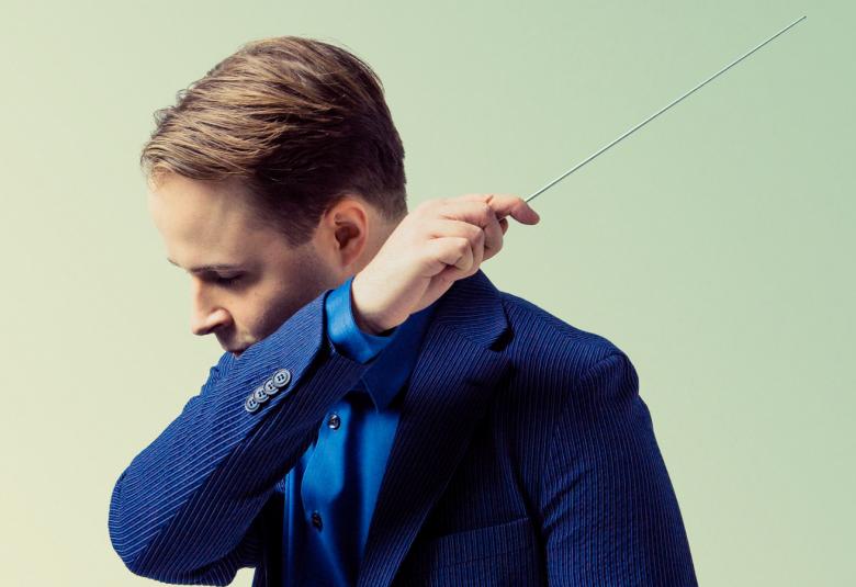 Kuvassa on Taavi Oramo johtamassa orkesteria ja hänestä näkyy vain ylävartalo. Oramo käsi edessä ja katse alas.