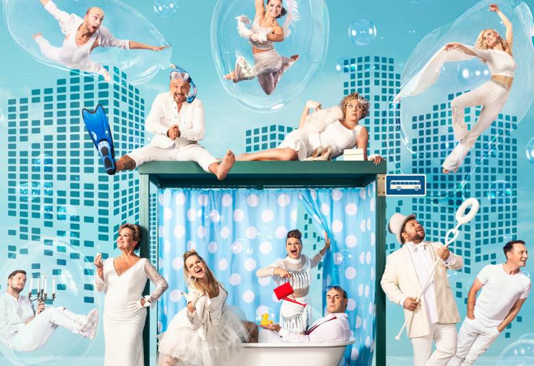 Kuvassa on UIT:n esiintyjiä valkoisissa asuissa vaaleansinisellä taustalla. Esiintyjät ovat kuvassa lentämässä taivaalla ja kylpyammeessa ja sen ympärillä.  Osa on tanssiliikkeessä.